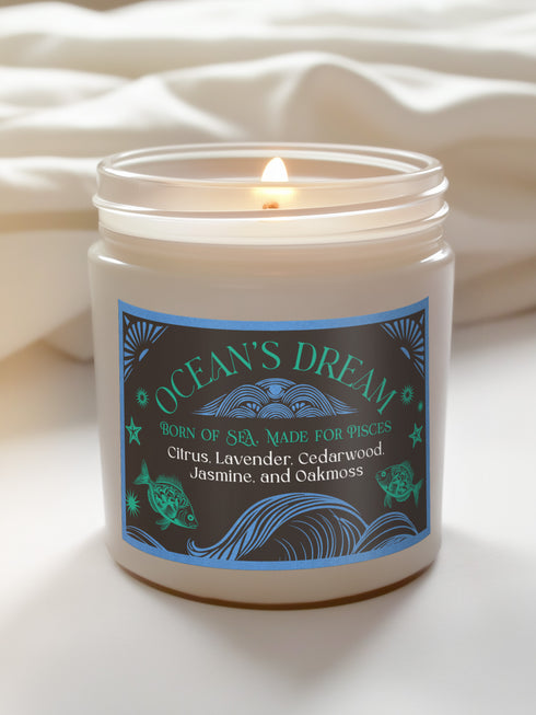 A 9 oz white soy candle in clear glass jar with blue and turquoise oceanic label featuring wave patterns and fish motifs, titled "Ocean's Dream" with a lit cotton wick.